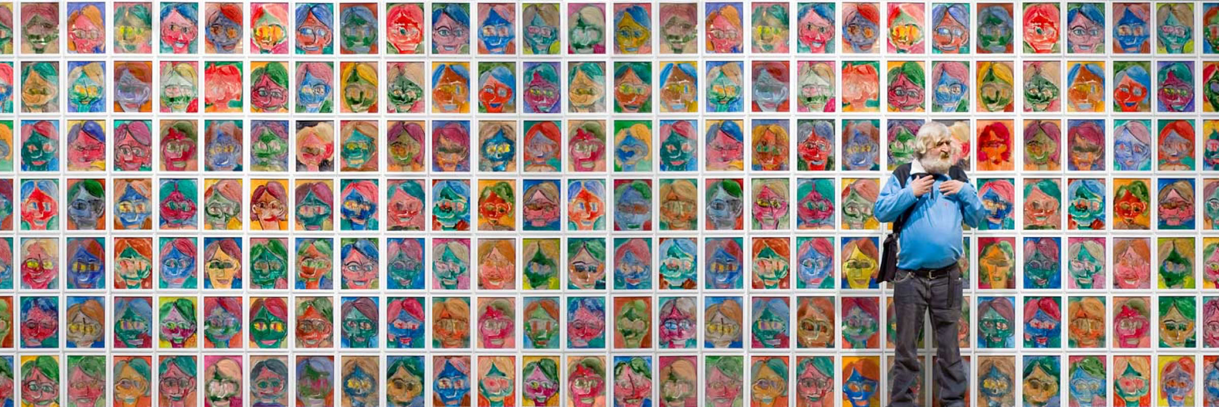 Auf dem Bild steht ein älterer Mann mit weißem Haar und Bart vor einer großen Wand voller bunter, expressiver Porträtgemälde. Es zeigt den Maler Stephan Kramer vor seinen Arbeiten. "Selbstportraits zur Dämonbändigung" betitelt er selbst diese Serie seines Oeuvres, die etwa 5000 Exponate umfasst. Die Porträts sind in vielen Reihen und Spalten angeordnet und zeigen stilisierte Gesichter in kräftigen Farben. 